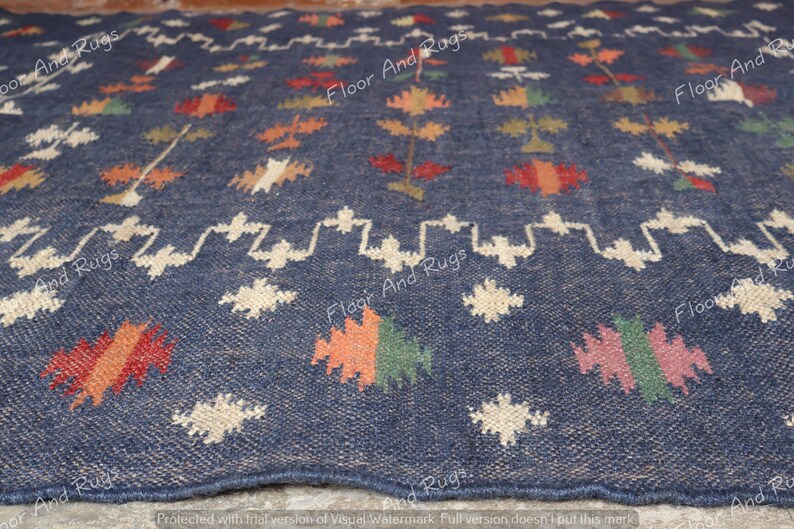 May include: A close-up of a blue rug with a white, orange, red, and green geometric pattern. The rug has a woven texture and is made of natural fibers.