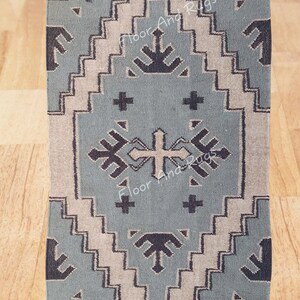 May include: A rectangular area rug featuring a dusty blue background with a large central diamond motif in beige and dark charcoal. The design includes geometric patterns, crosses, and arrow-like shapes. The rug has fringed ends and text reading Floor And Rugs.