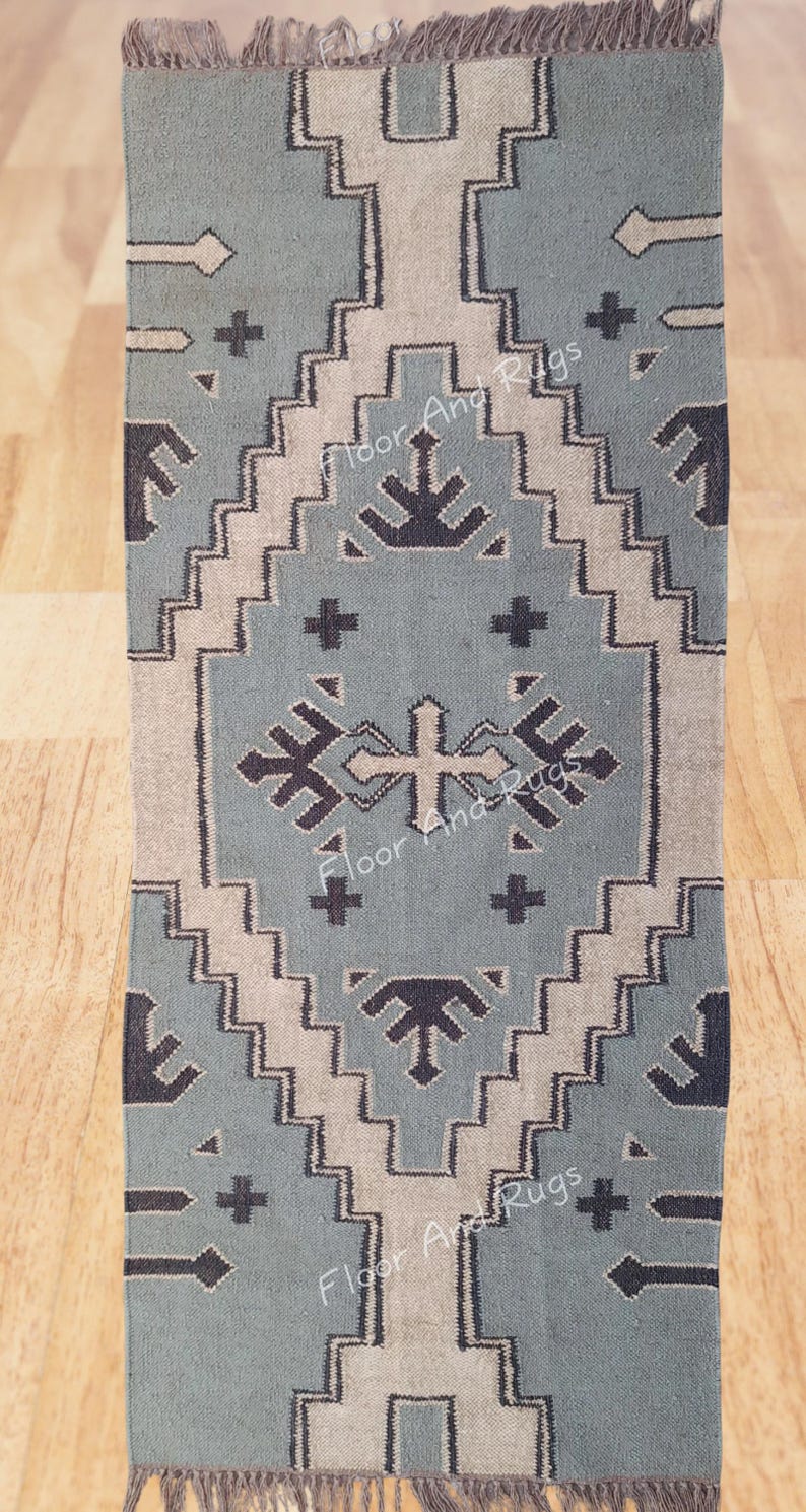 May include: A rectangular area rug featuring a dusty blue background with a large central diamond motif in beige and dark charcoal. The design includes geometric patterns, crosses, and arrow-like shapes. The rug has fringed ends and text reading Floor And Rugs.