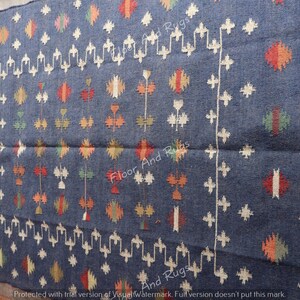 May include: A blue wool rug with a geometric pattern in white, red, orange, and green. The rug has fringe on all four sides.