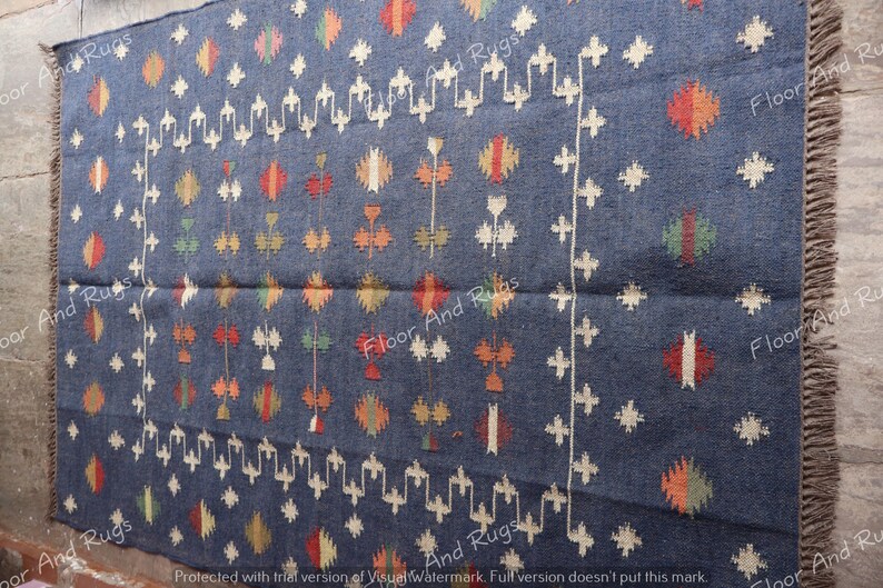 May include: A blue wool rug with a geometric pattern in white, red, orange, and green. The rug has fringe on all four sides.