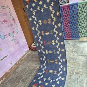 May include: A long, rectangular blue runner rug featuring a repeating geometric pattern in cream, red, green, and yellow. The rug has a textured, woven appearance with a fringed edge at the bottom, showcasing a symmetrical design of small motifs.