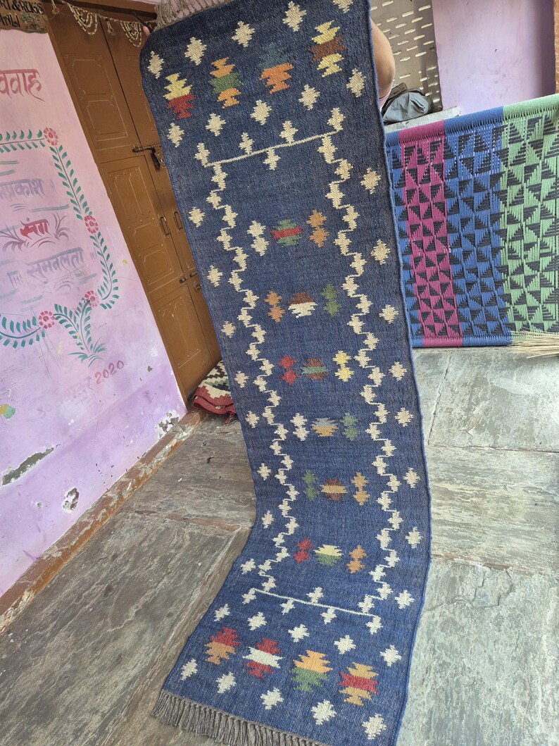 May include: A long, rectangular blue runner rug featuring a repeating geometric pattern in cream, red, green, and yellow. The rug has a textured, woven appearance with a fringed edge at the bottom, showcasing a symmetrical design of small motifs.
