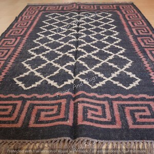 May include: A rectangular area rug featuring a dark charcoal background with a central white diamond lattice pattern. The border displays a pink Greek key motif. The rug is finished with short, light-colored fringe along the bottom edge.