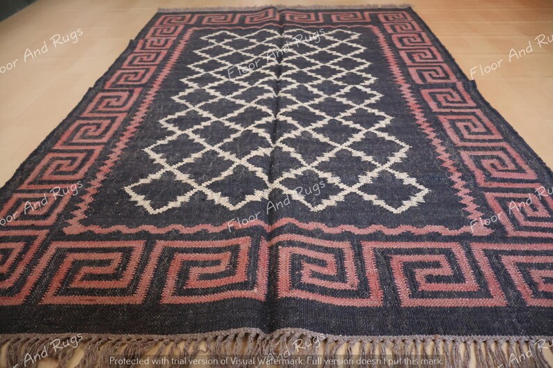May include: A rectangular area rug featuring a dark charcoal background with a central white diamond lattice pattern. The border displays a pink Greek key motif. The rug is finished with short, light-colored fringe along the bottom edge.