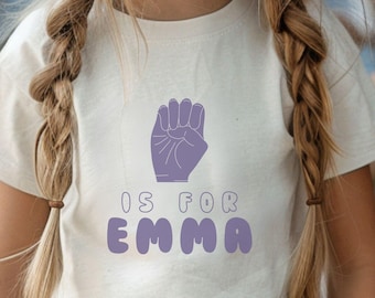 May include: A cream-coloured t-shirt featuring a dark purple graphic. The graphic shows a hand in sign language and the text "IS FOR EMMA" in purple bubble letters. The shirt is designed for children.