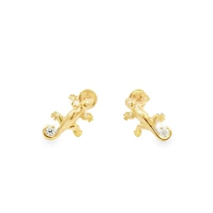 May include: A pair of gold-colored lizard stud earrings. Each earring features a small, clear gemstone near the tail. The lizards are detailed with tiny legs, a head, and a curled tail. The earrings are set against a white background.