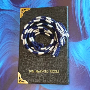 May include: A black leather-bound journal with gold accents and the inscription "TOM MARVOLO RIDDLE" on the front cover. A blue and white striped cord with tassels is resting on the cover.