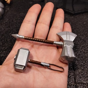 May include: Two silver-colored toy weapons, a hammer and an axe, with brown handles. The weapons are held in a hand.