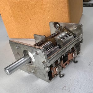 Puede incluir: Un condensador variable plateado vintage con múltiples placas metálicas y un eje cilíndrico largo. El dispositivo tiene un marco rectangular y componentes de color cobre. Una caja marrón texturizada está en el fondo.
