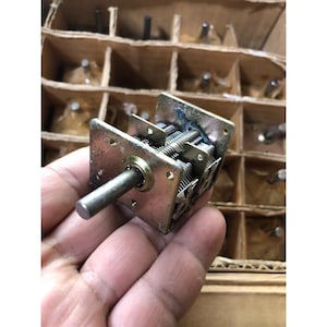 Puede incluir: Un condensador variable vintage con un marco de metal y múltiples placas internas. El condensador tiene un eje central y se sostiene en una mano. El fondo muestra una caja de cartón con múltiples compartimentos.