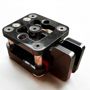 Puede incluir: Un dispositivo mecánico negro con una parte superior cuadrada que presenta múltiples recortes circulares y seis tornillos plateados. El dispositivo tiene dos paneles laterales negros brillantes y soportes de color cobre.