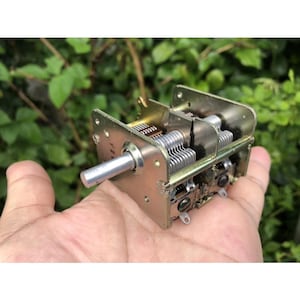 Puede incluir: Un condensador variable metálico vintage con un eje plateado y múltiples placas paralelas. El dispositivo rectangular tiene un acabado dorado y se sostiene en una mano. El fondo es verde borroso.
