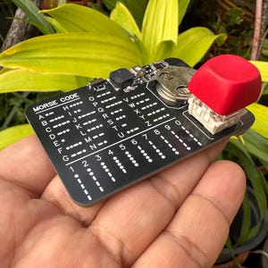 Op de afbeelding: Een zwarte printplaat met de woorden "MORSE CODE" en morsecode tekens in het wit gedrukt. Een grote rode vierkante knop is aan de printplaat bevestigd. De printplaat wordt in een hand gehouden tegen een achtergrond van groen gebladerte.