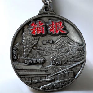 Puede incluir: Un llavero redondo de color plateado con un diseño en relieve detallado. El colgante presenta texto japonés en rojo, un paisaje montañoso, edificios y un teleférico. El diseño incluye el texto "Hakone".