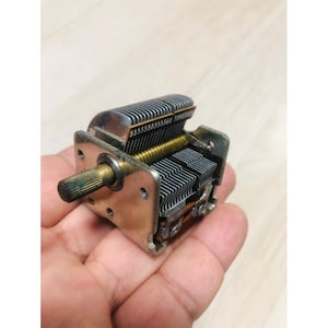 Puede incluir: Un condensador variable plateado y dorado vintage, un componente utilizado en circuitos de radio. El dispositivo presenta múltiples placas metálicas paralelas y un eje giratorio. El componente se sostiene en una mano.