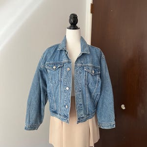 May include: A light blue denim jacket with silver buttons and two chest pockets. The jacket is displayed on a mannequin with a beige skirt. The jacket has a classic collar and long sleeves.