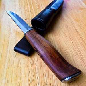 May include: A handcrafted knife with a wooden handle and a stainless steel blade, accompanied by a black leather sheath. The knife is displayed on a wooden surface, showcasing its polished finish and detailed craftsmanship.