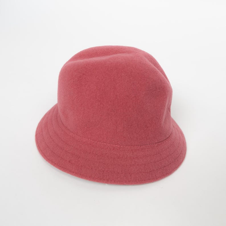 May include: A solid dusty rose bucket hat with a rounded crown and a downward-sloping brim. The hat features a soft, felted texture throughout its surface, with visible stitching details along the brim and a simple, unadorned design.