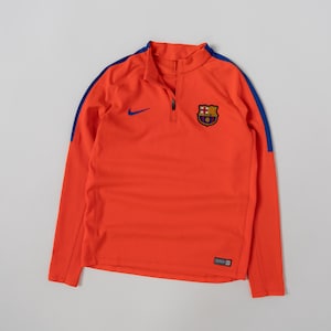May include: A bright orange long-sleeve quarter-zip pullover featuring a blue Nike swoosh logo on the right chest and the FC Barcelona football club crest on the left chest. The garment includes blue shoulder stripes and a small black label at the hem.
