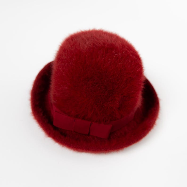 May include: A deep red fuzzy hat with a rounded crown and a short, curved brim. The hat features a matching red ribbon band wrapped around the base of the crown, which is adorned with a small, flat bow detail on the front side of the hat.