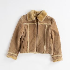 May include: A tan suede jacket featuring a plush, light-colored faux fur collar and matching faux fur trim along the vertical front panels and sleeve cuffs. The jacket includes a central zipper closure and long sleeves with a soft, textured finish.