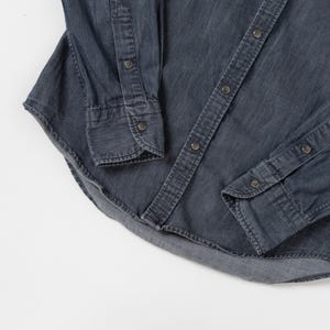 May include: A long-sleeved button-down shirt in a dark charcoal gray denim fabric. The shirt features a front placket with metallic snap buttons, matching snap buttons on the cuffs, and a curved hemline with visible stitching along the edges.