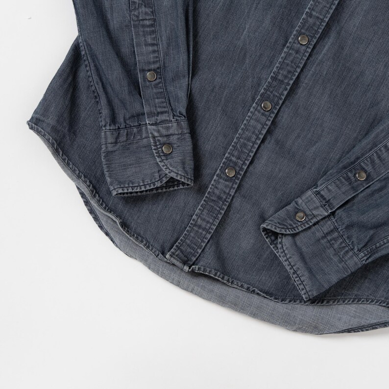 May include: A long-sleeved button-down shirt in a dark charcoal gray denim fabric. The shirt features a front placket with metallic snap buttons, matching snap buttons on the cuffs, and a curved hemline with visible stitching along the edges.