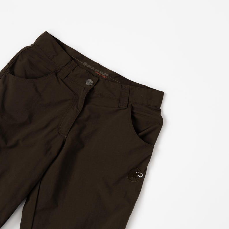 Puede incluir: Primer plano de unos pantalones marrones para actividades al aire libre con dos bolsillos delanteros y cierre de bot&oacute;n. Los pantalones tienen un peque&ntilde;o logotipo en el bolsillo derecho y la marca es visible en la cintura. Los pantalones son de material ligero.