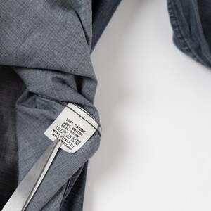 May include: A dark gray cotton shirt with a white interior care label that reads 100% COTONE, 100% COTTON, 100% COTON, and includes wash instructions. A pair of silver metal scissors is positioned at the bottom edge of the fabric label.
