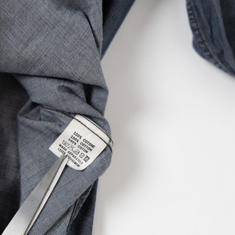 May include: A dark gray cotton shirt with a white interior care label that reads 100% COTONE, 100% COTTON, 100% COTON, and includes wash instructions. A pair of silver metal scissors is positioned at the bottom edge of the fabric label.