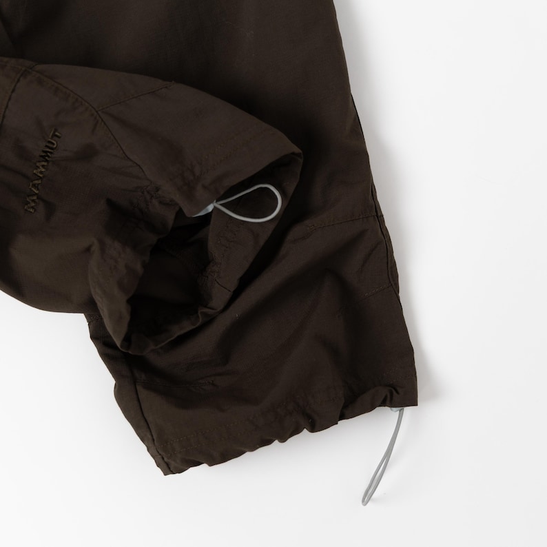Puede incluir: Primer plano de un pantal&oacute;n marr&oacute;n oscuro con un cord&oacute;n en el tobillo. El pantal&oacute;n tiene una textura ligeramente brillante y la marca "MAMMUT" es visible en el lateral. El cord&oacute;n es de color gris claro.