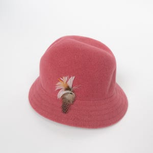 May include: A dusty rose felt bucket hat featuring a rounded crown and a downward-sloping brim. The hat is adorned with a decorative accent consisting of a cluster of white, orange, and brown feathers attached to the side of the crown.