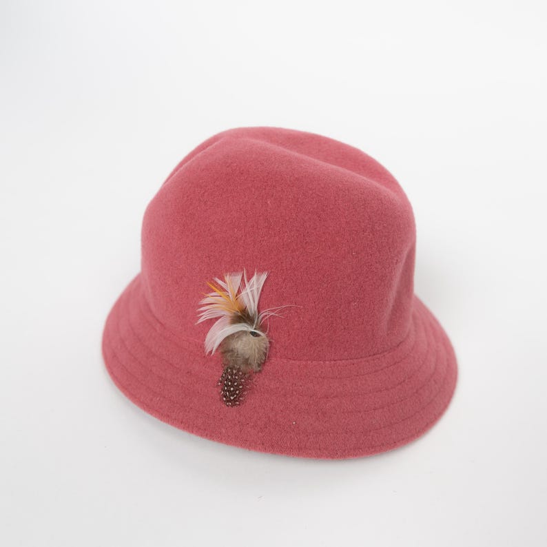 May include: A dusty rose felt bucket hat featuring a rounded crown and a downward-sloping brim. The hat is adorned with a decorative accent consisting of a cluster of white, orange, and brown feathers attached to the side of the crown.
