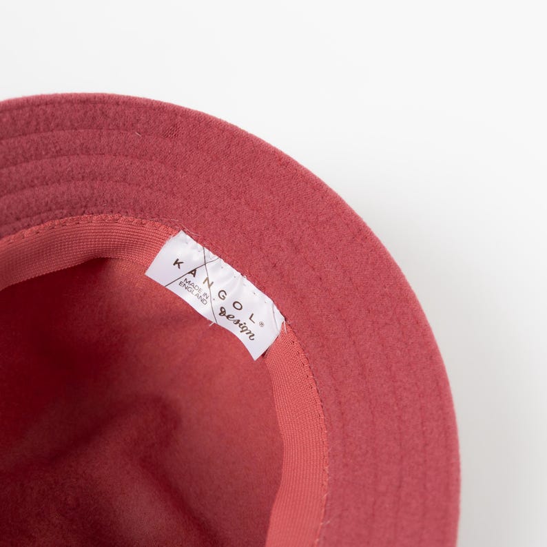 May include: A dusty rose felt bucket hat with a wide brim and a matching interior sweatband. A white fabric label is sewn into the inner rim, featuring the black text KANGOL, MADE IN ENGLAND, and design in a cursive script font.