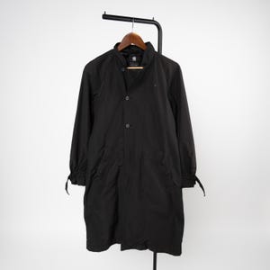 May include: A black long-length coat featuring a high collar, a front button closure, and long sleeves with adjustable tie cuffs. The garment includes two front pockets and a G-Star Raw brand label visible on the interior neck area.