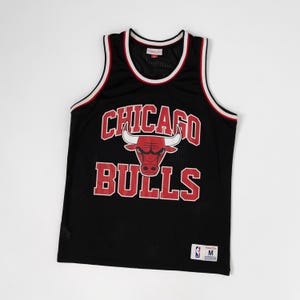 Puede incluir: Una camiseta de baloncesto negra sin mangas con el texto CHICAGO BULLS en rojo con borde blanco y un gráfico de cabeza de toro roja en el centro. El cuello y las sisas tienen ribetes blancos y rojos. Una etiqueta blanca con la letra M está abajo.
