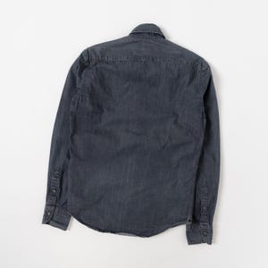 May include: A long-sleeved button-down shirt in a dark charcoal gray color. The shirt features a classic collar, a back yoke, and cuffs with buttons. The fabric has a subtle texture and a slightly faded appearance, with visible stitching along the seams.