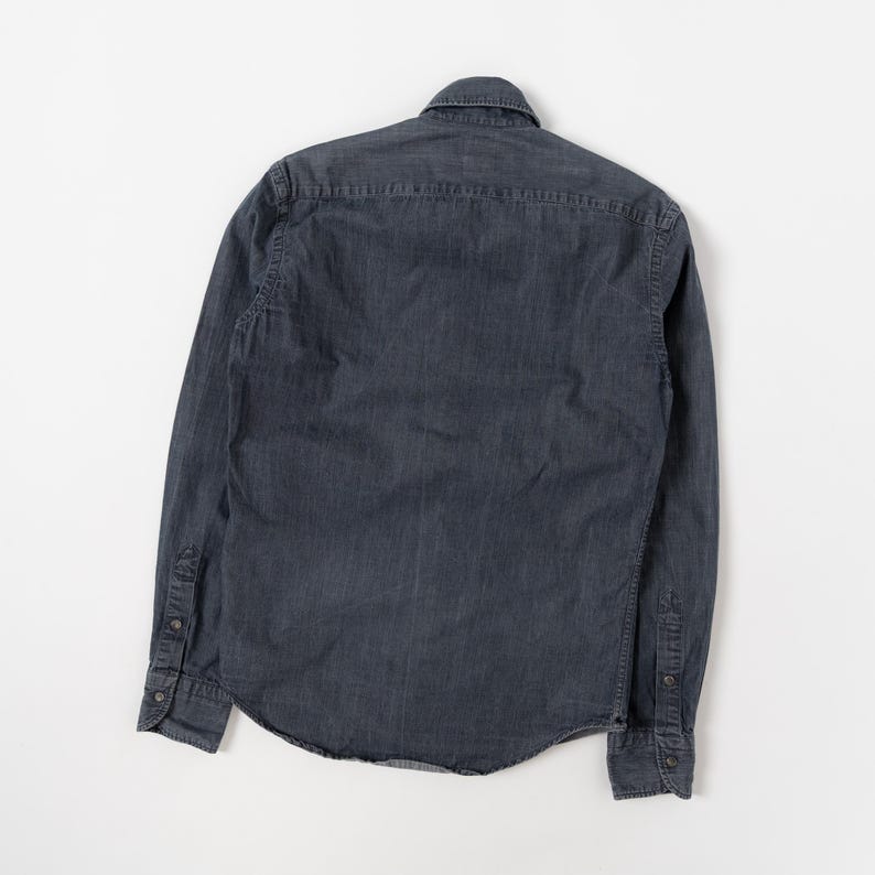 May include: A long-sleeved button-down shirt in a dark charcoal gray color. The shirt features a classic collar, a back yoke, and cuffs with buttons. The fabric has a subtle texture and a slightly faded appearance, with visible stitching along the seams.