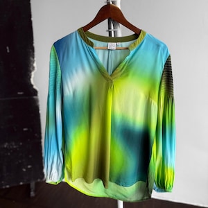 Könnte beinhalten: Eine langärmlige Bluse mit V-Ausschnitt und einem lebendigen Batikmuster in Blau-, Grün- und Gelbtönen. Die Ärmel haben eine geraffte Textur an Schultern und Manschetten. Die Bluse hängt an einem Holzkleiderbügel.