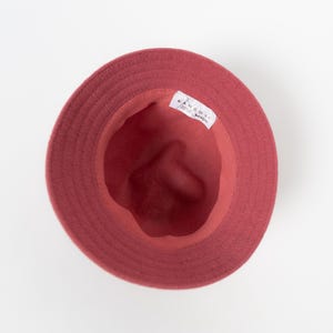 May include: A top-down view of a solid red bucket hat with a circular brim. The interior of the hat is lined with a matching red fabric and features a white rectangular label with the black text KANGOL printed on it, positioned along the inner rim.