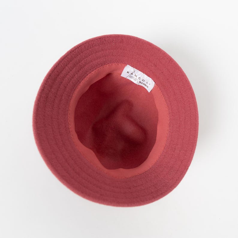 May include: A top-down view of a solid red bucket hat with a circular brim. The interior of the hat is lined with a matching red fabric and features a white rectangular label with the black text KANGOL printed on it, positioned along the inner rim.