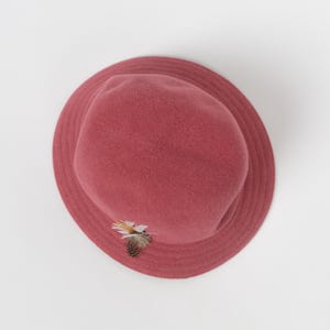 May include: A dusty rose-colored felt hat with a rounded crown and a wide brim. The hat features a small decorative cluster of natural-toned feathers attached to the side, providing a subtle textural contrast to the smooth, matte felt surface.