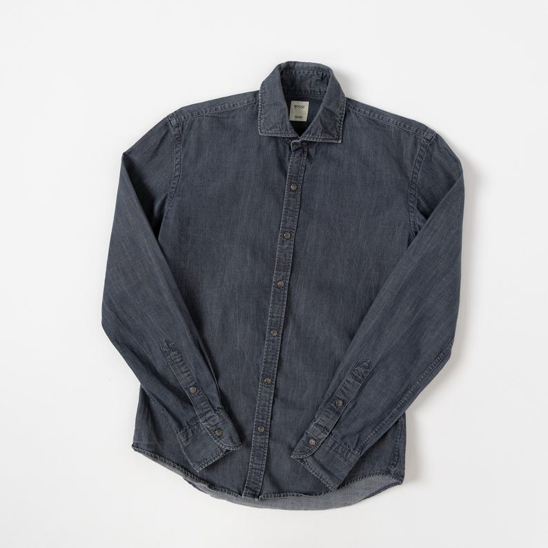 May include: A long-sleeve button-down shirt in a dark charcoal gray denim fabric. The shirt features a standard collar, a front button placket with matching dark buttons, and buttoned cuffs on the sleeves. A small white label is visible at the neck.