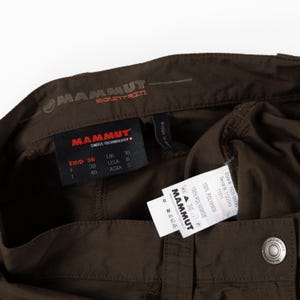 Puede incluir: Primer plano de pantalones marrones para exteriores con varias etiquetas. Las etiquetas muestran la marca "MAMMUT" e informaci&oacute;n de tallas. Los pantalones est&aacute;n hechos de 100% poliamida. Los pantalones tienen cierre de bot&oacute;n y costura delantera.