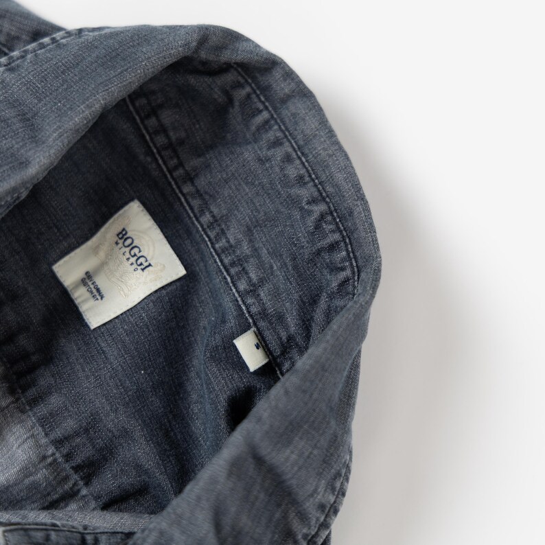 May include: A dark gray button-down shirt with a textured fabric finish. The interior collar features a white fabric label with the text Boggi Milano and Easy Formal Cotton Fit, along with a small white size tag displaying the letter S.