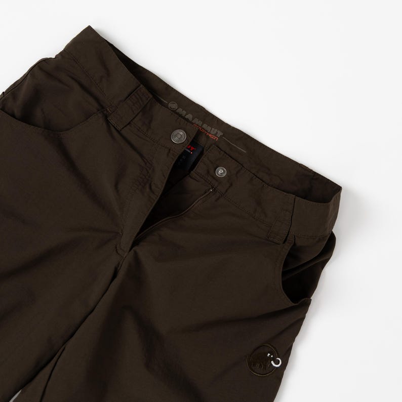 Puede incluir: Pantalones de exterior marr&oacute;n oscuro con cierre de bot&oacute;n y cremallera. Los pantalones tienen dos bolsillos delanteros y un peque&ntilde;o parche con el logotipo cerca del bolsillo derecho. La marca "Mammut" es visible en la cintura.