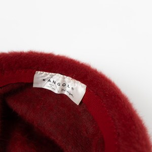 May include: A deep red fuzzy hat with a soft, textured surface. Inside the hat, a white fabric label is stitched to the inner band, featuring the black text KANGOL, the word design, and the phrase MADE IN ENGLAND printed in a clear font.