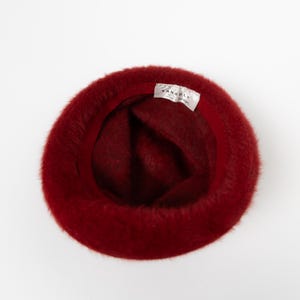 May include: A deep red fuzzy bucket hat with a soft, textured exterior. The interior features a dark red fabric lining and a white rectangular tag sewn into the inner rim with the black text KANGOL and the brand logo clearly visible.
