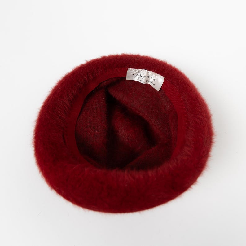 May include: A deep red fuzzy bucket hat with a soft, textured exterior. The interior features a dark red fabric lining and a white rectangular tag sewn into the inner rim with the black text KANGOL and the brand logo clearly visible.